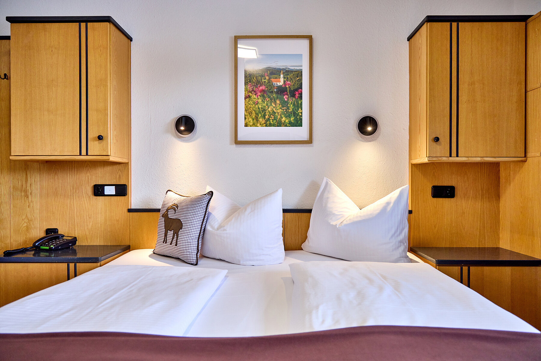 Detailaufnahme des Doppelbetts, das beidseitig von Hängeschränken eingefasst ist. Darüber befinden sich zwei Leuchten und ein Landschaftsbild. 