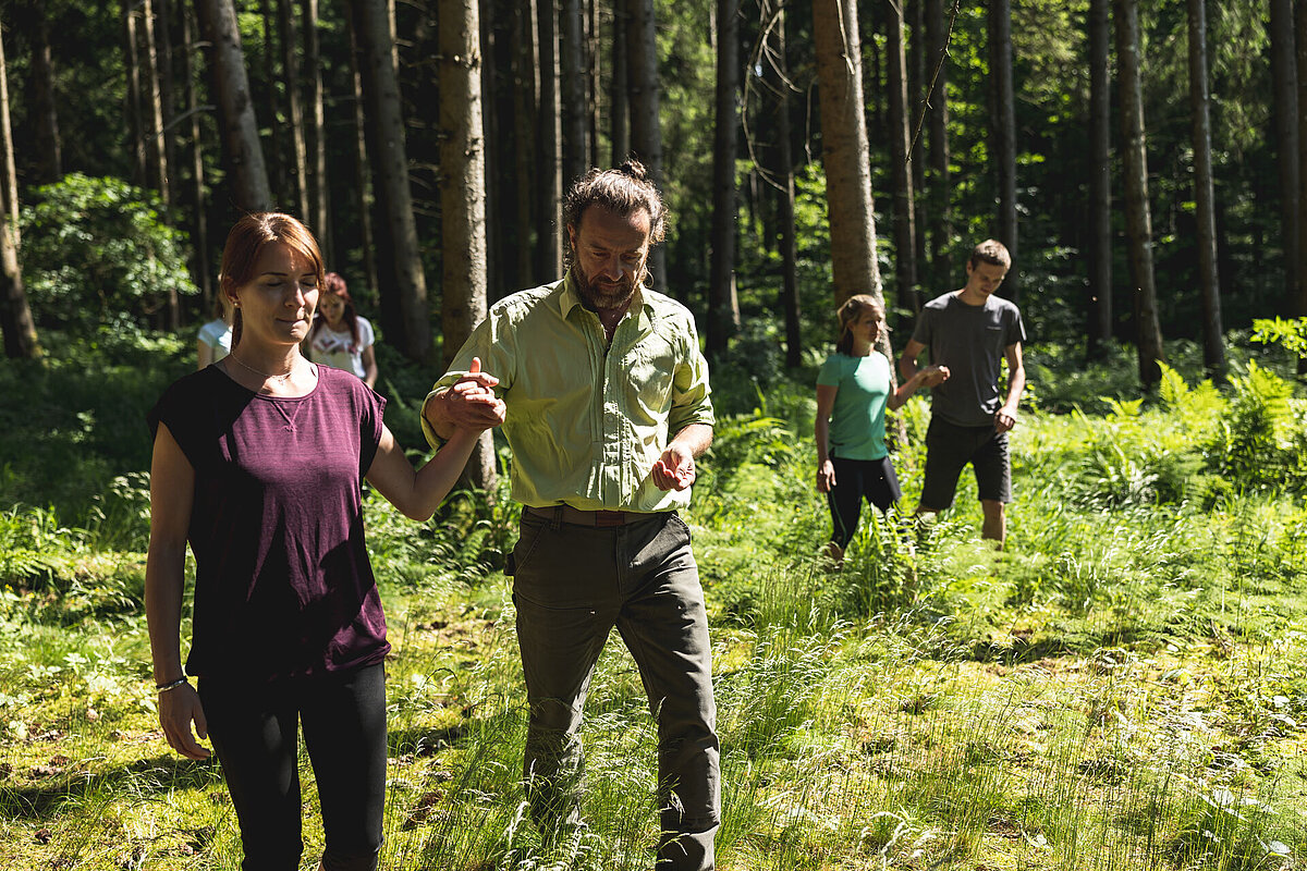 Personen in Zweiergruppen schreiten durch einen Wald . dabei hat jeweils eine Person die Augen geschlossen, um die Umgebung mit anderen Sinnen zu erleben.