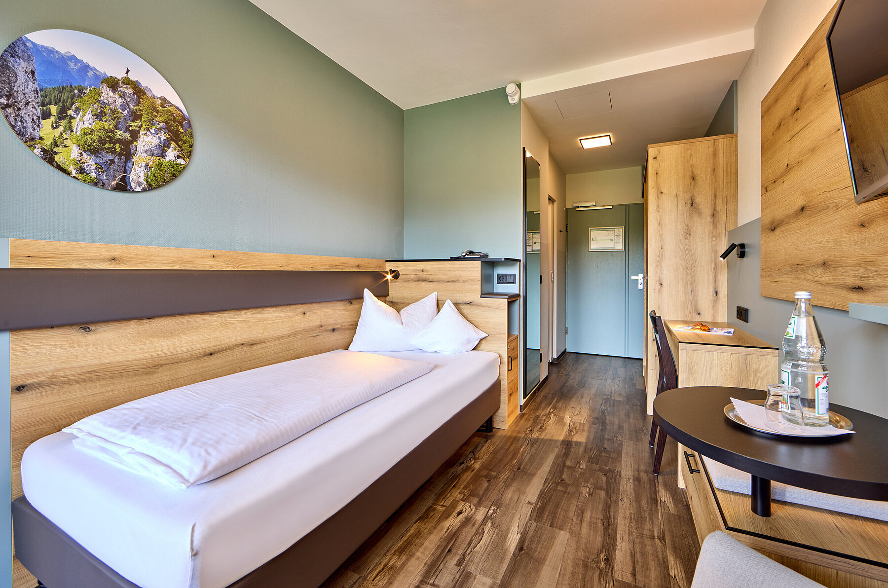 Das weiß bezogenen Bett steht an einer hellen Holzwand mit Lederapplikation - darüber ist ein hübsches rundes Bild mit heimischen Bergmotiv. 