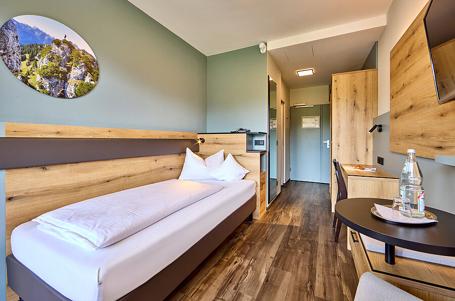 Das weiß bezogenen Bett steht an einer hellen Holzwand mit Lederapplikation - darüber ist ein hübsches rundes Bild mit heimischen Bergmotiv. 