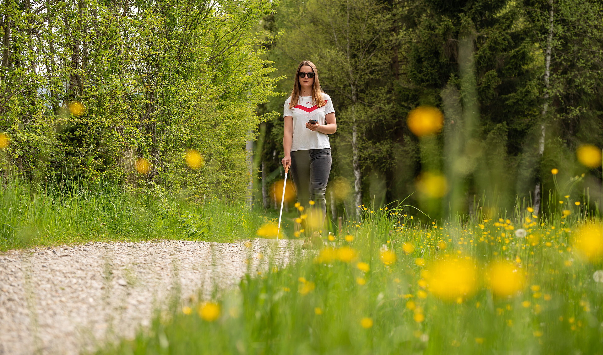 Eine blinde Frau läuft mit dem Taststock über einen Schotterweg im Grünen.