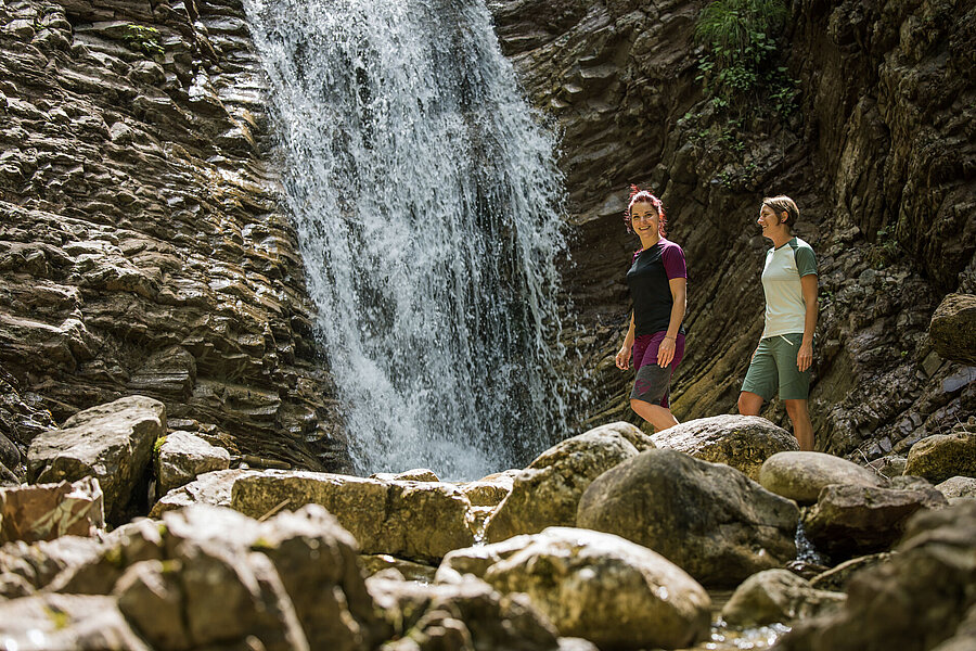 Zwei Wanderinnen balancieren für große Steine bei einem Wasserfall.