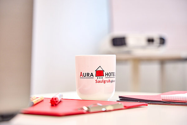 Auf dem Tisch steht eine Tasse mit AURA-Logo und liegt Schreibmaterial bereit.