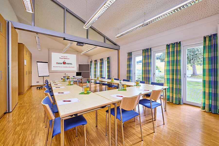 Der helle Tagungsraum ist mit einer Blockbestuhlung ausgestattet. Durch die blau-gelb karierten Vorhänge an den bodentiefen Fenstern wirkt der Raum freundlich.