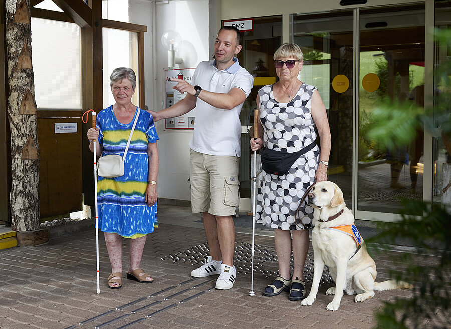 Der Teamleiter der Gästebetreuung beschreibt zwei Urlauberinnen mit Führhund die Wegführung vor dem Hoteleingang.