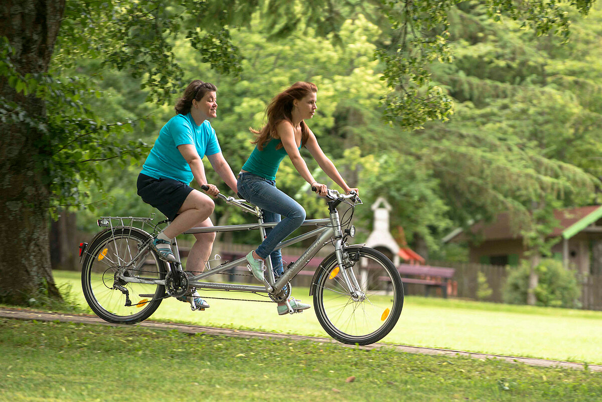 Zwei junge Frauen radeln auf einem Tandem durch den Hotelpark.