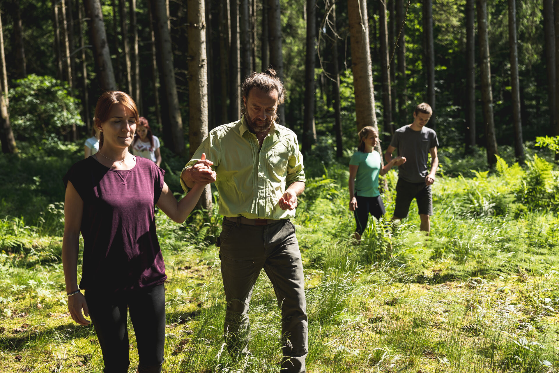 Mehrere Personen führen sich gegenseitig durch den Wald, dabei hat eine immer die Augen geschlossen.