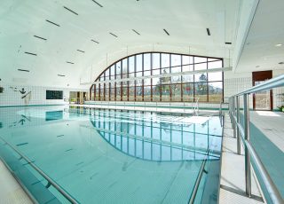 Großes türkises Schwimmbecken in Halle mit großer Fensterfront zum Bergpanorama, Link zu Schwimmbad und Sauna
