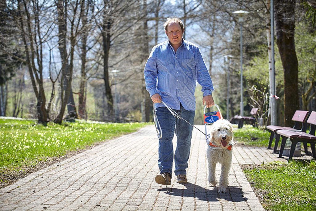Ein Mann läuft einen Parkweg entlang. Er hält seinen Hund am Halsband und einem speziellen Griff für Blinde.
