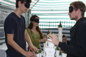 Zwei Männer und eine junge Frau tragen Augenbinden und ertasten ein Modell von Neuschwanstein