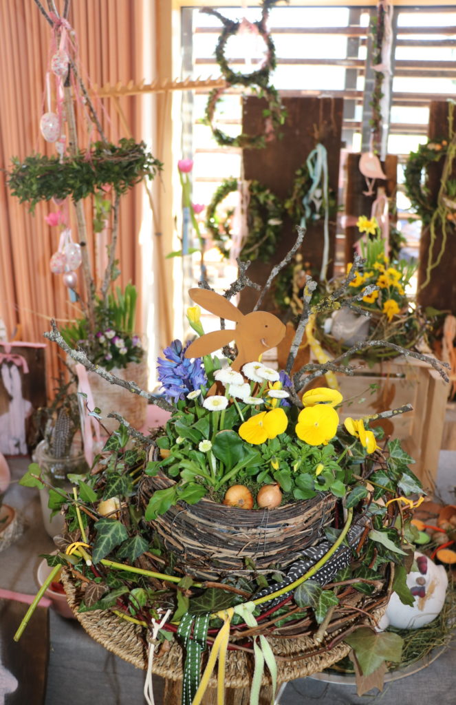 Ein Weidenkorb mit bunt bepflanzten Frühlingsblumen und einem Metallhasen bildet den Mittelpunkt des Fotos. Im Hintergrund sind verschiedene Floristikartikel wie Kränze und Gestecke zu sehen.