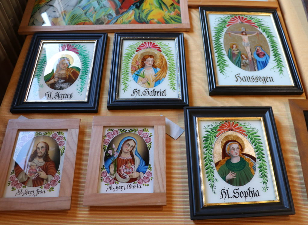 Am Stand werden Hinterglasmalereien angeboten. Fünf kleinere Bilder zeigen Porträts von Heiligen, ein größeres ist ein Haussegen und zeigt die Kreuzigungsszene Jesu.