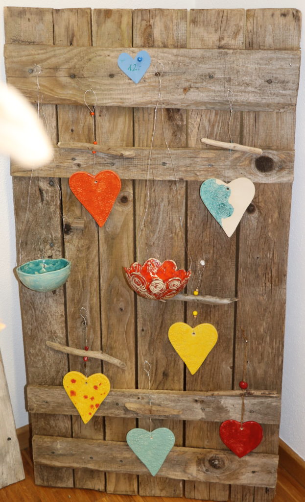 Auf einem alten Holzfensterladen sind bunte, getöpferte Herzen und kleine Hängeschalen dekoriert.
