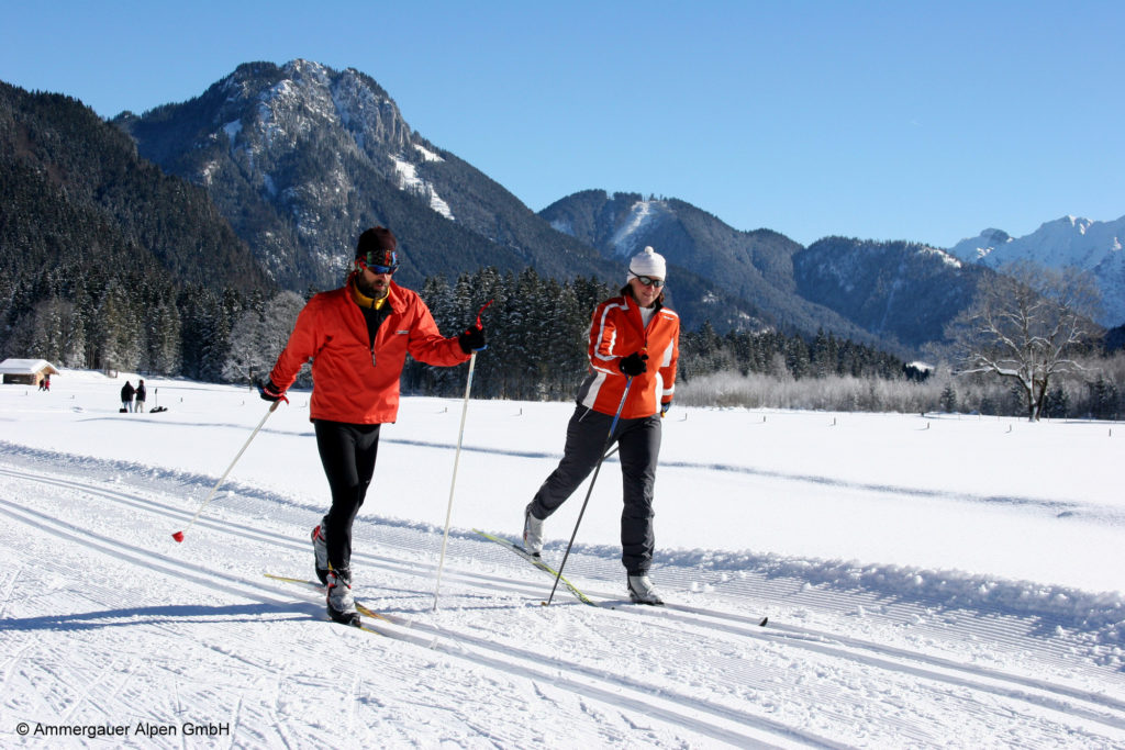 Eine Frau und ein Mann bewegen sich auf Langlaufskiern durch die winterliche Landschaft. Im Hintergrund sind die Berge zu sehen.
