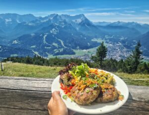 Vor der grandiosen Kulisse des Wettersteingebirges wird ein Teller mit frischem knackigen Salat und Kaspressknödeln hochgehalten.