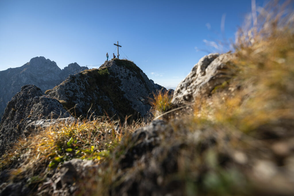 Panoramaaufnahme eines Berggipfels, an dessen Kreuz ein Wanderer steht