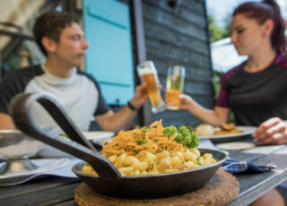 Im Vordergrund ist eine Pfanne voller Käsespätzle und ein Paar prostet sich zu