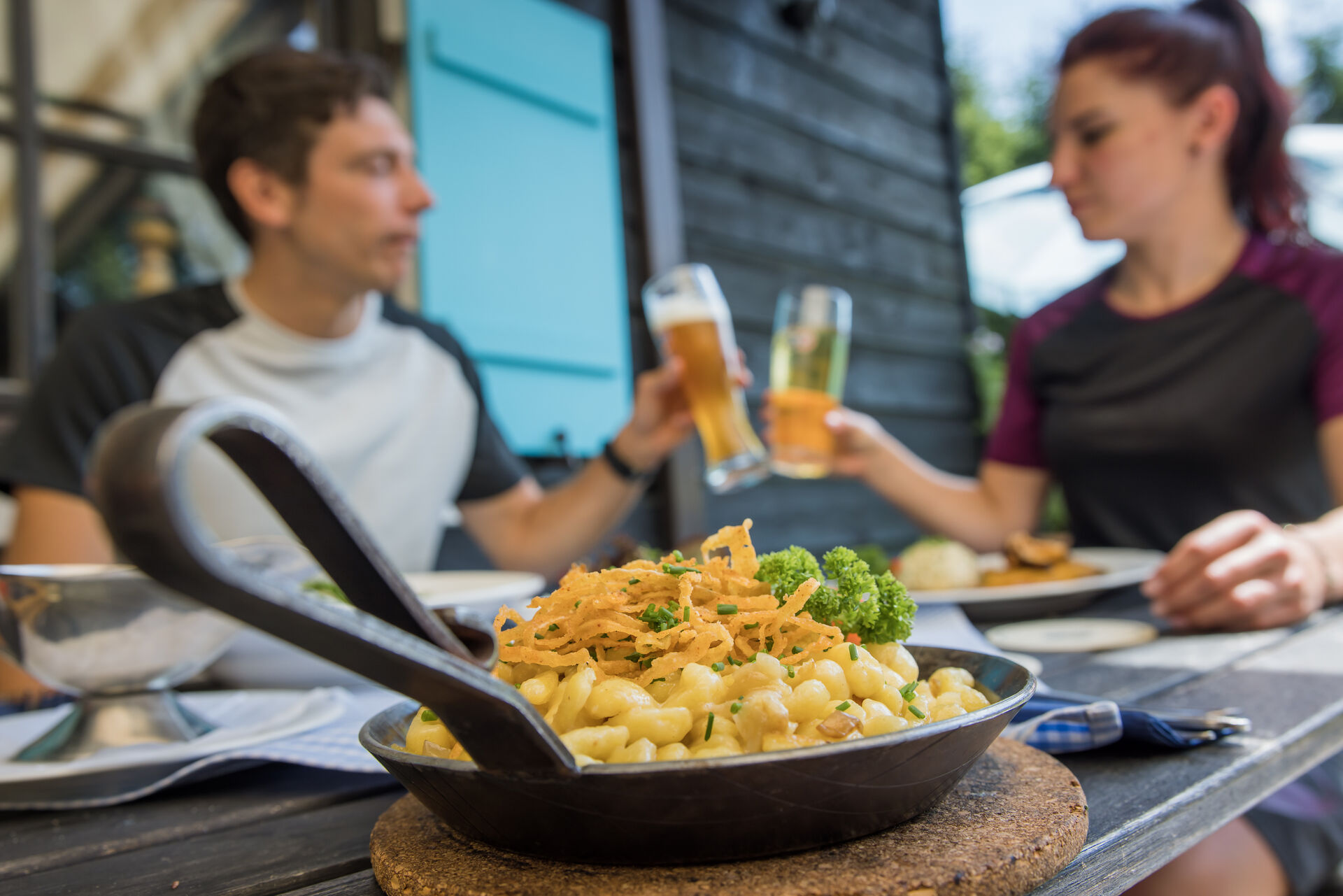 Im Vordergrund ist eine Pfanne voller Käsespätzle und ein Paar prostet sich zu